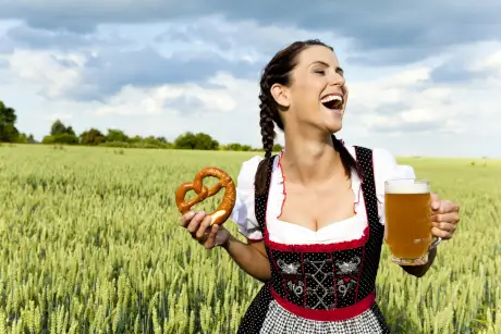 Fröhliche Frau im Dirndl hält eine Brezel und ein Bier vor einem Getreidefeld – ein Motiv für „Im OB ist immer Oktoberfest“.},